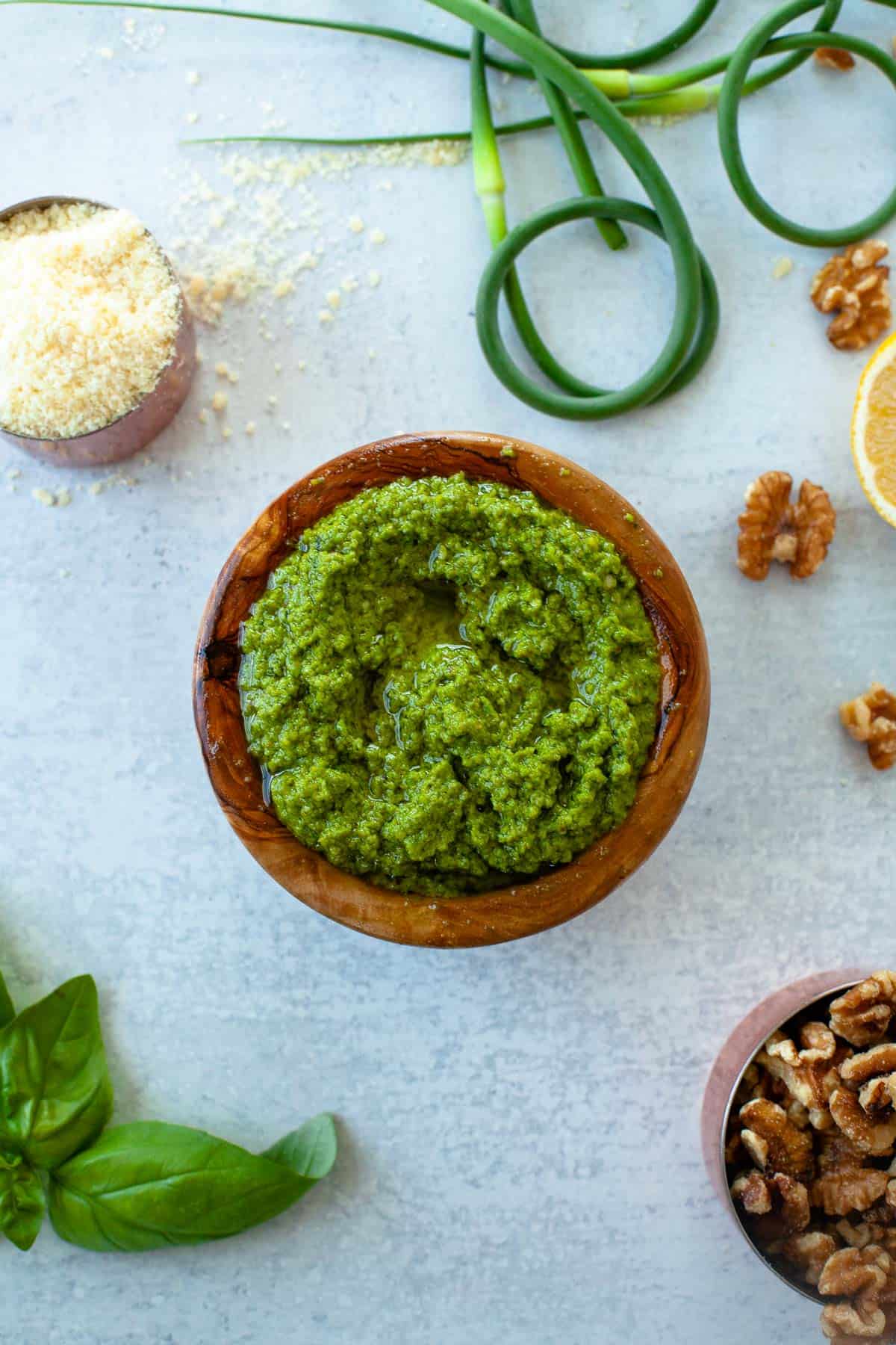 pesto in small wooden bowl. cup of parmesan. scapes. cup of walnuts. basil. lemon slice. on grey counter. 