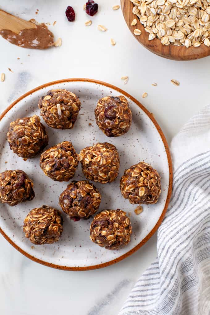 Almond butter protein bites on a white speckled plate with an orange rim. A bowl of rolled oats and spoon with almond butter in corner