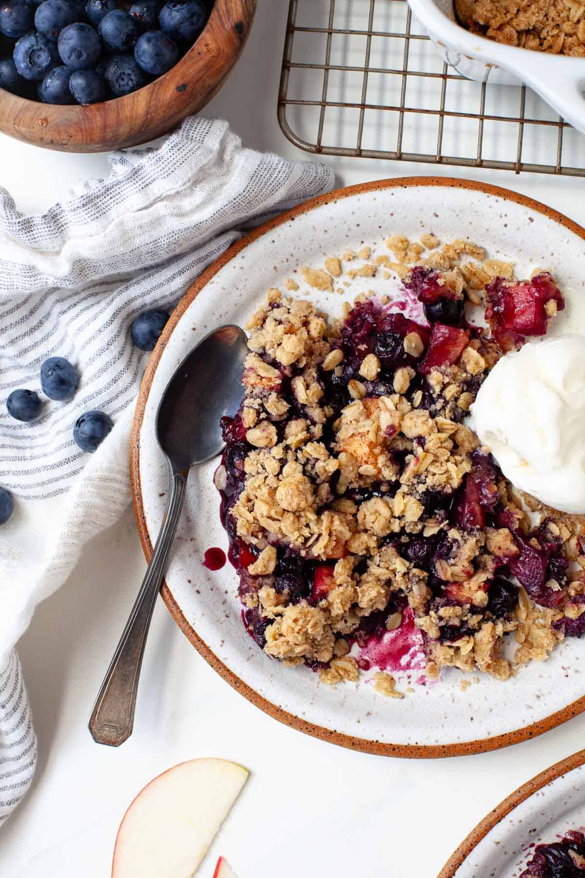 Plate of blueberry apple crisp with scoop of vanilla ice cream beside kitchen towel and bowl of blueberries